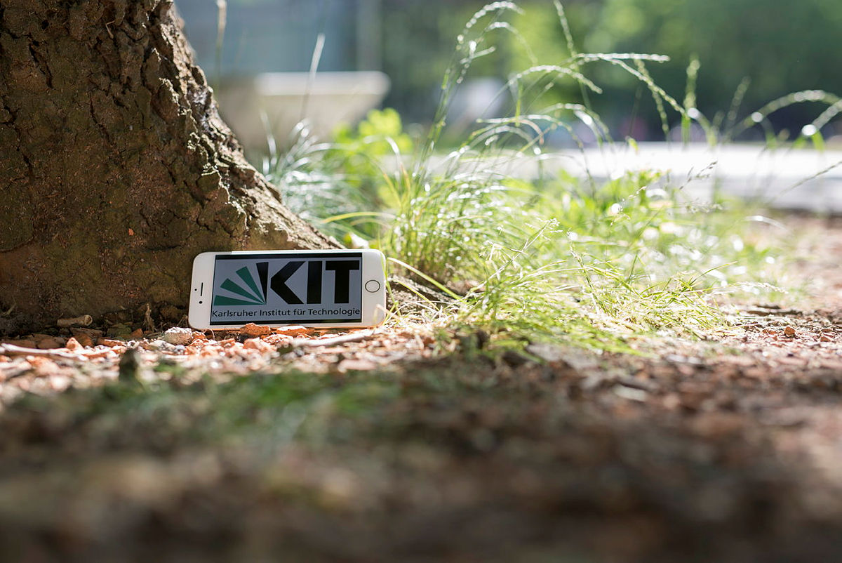 Ein Handy liegt auf dem Boden unter einem Baum. Der Bildschirm des Handys zeigt das KIT Logo.