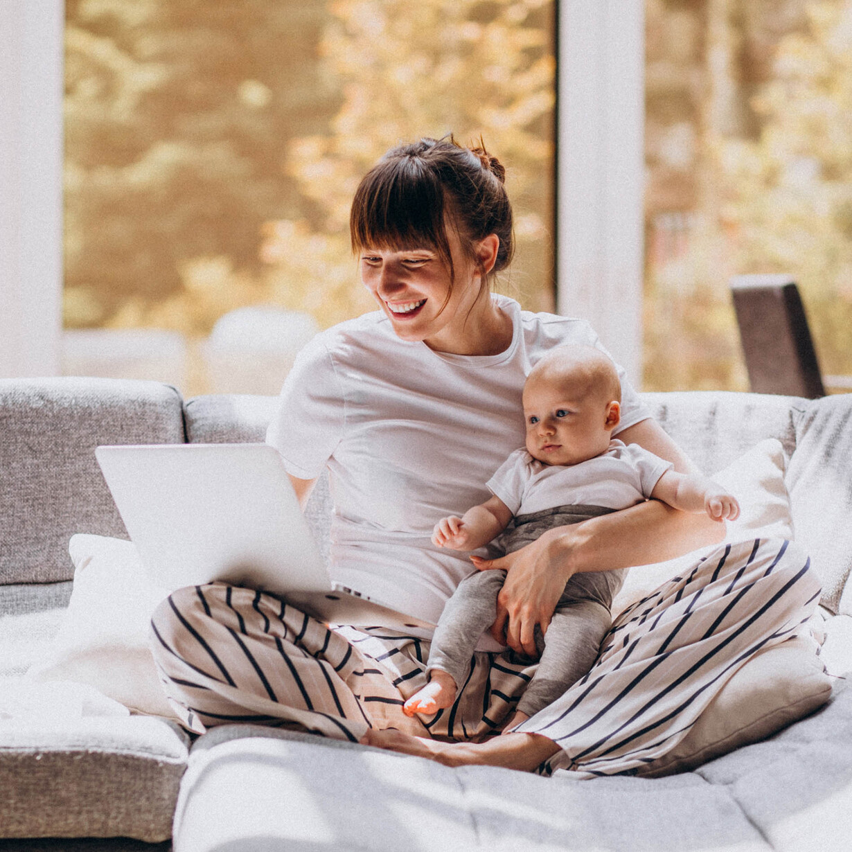 Eine Frau sitzt miz einem Baby im Arm auf dem Sofa und arbeitet an einem Laptop